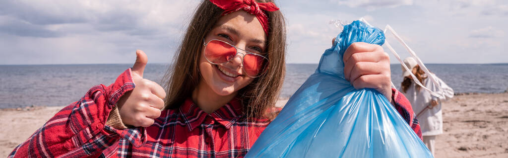 šťastná mladá žena ve slunečních brýlích držící pytel na odpadky a ukazující palec nahoru, prapor - Fotografie, Obrázek