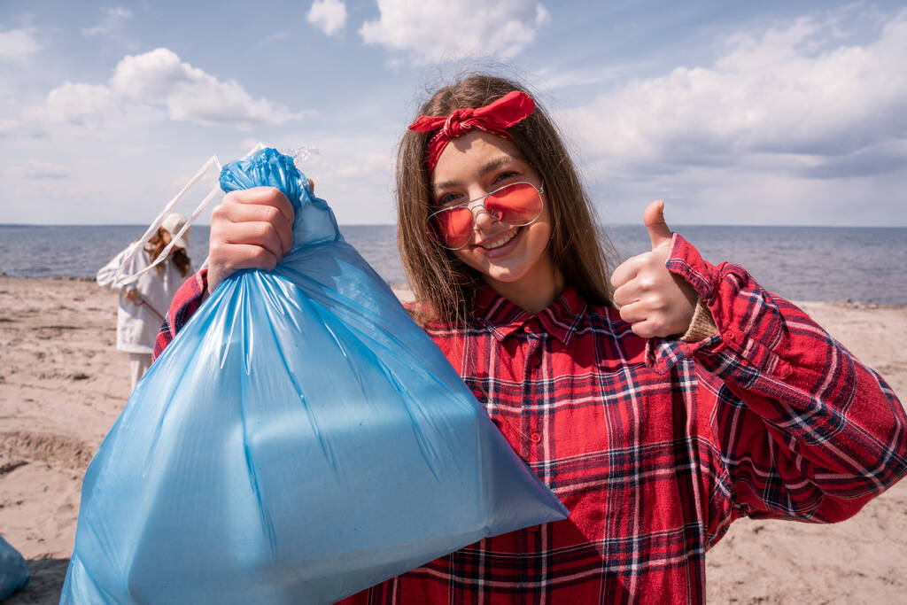 felice giovane donna in occhiali da sole tenendo sacchetto della spazzatura e mostrando pollice in su, concetto di ecologia  - Foto, immagini