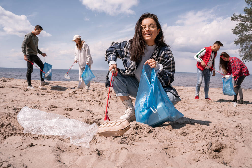 heureuse jeune femme ramassant des ordures avec grabber près du groupe de bénévoles  - Photo, image