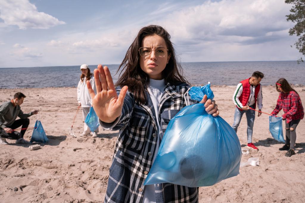 mujer joven mostrando gesto de stop cerca de grupo borroso de voluntarios recogiendo basura, concepto de ecología  - Foto, imagen