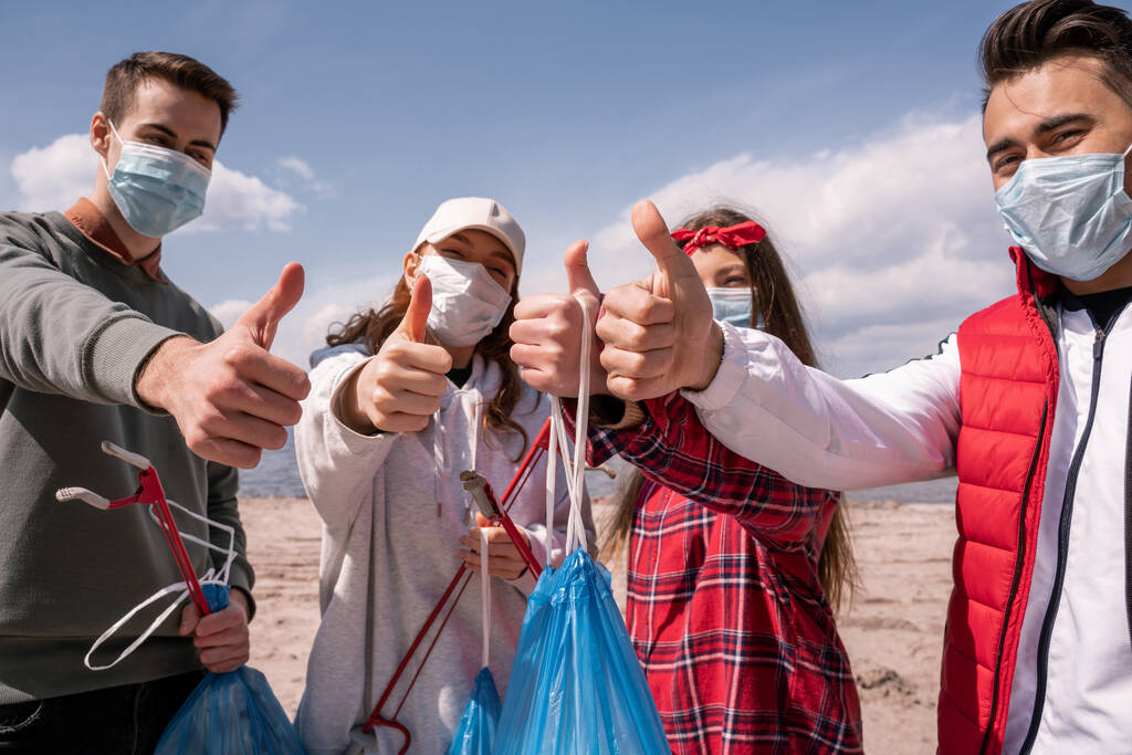 młodzi ochotnicy w maskach medycznych trzymający worki na śmieci i łapacze, podnoszący kciuki, podnoszący śmieciowy pomysł  - Zdjęcie, obraz
