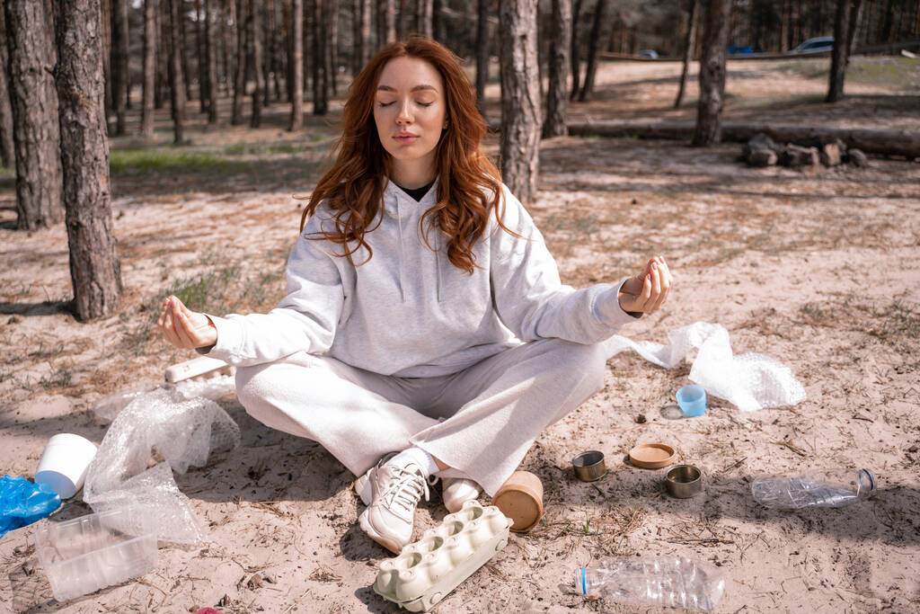 młoda kobieta z zamkniętymi oczami medytująca w pobliżu śmieci na ziemi - Zdjęcie, obraz