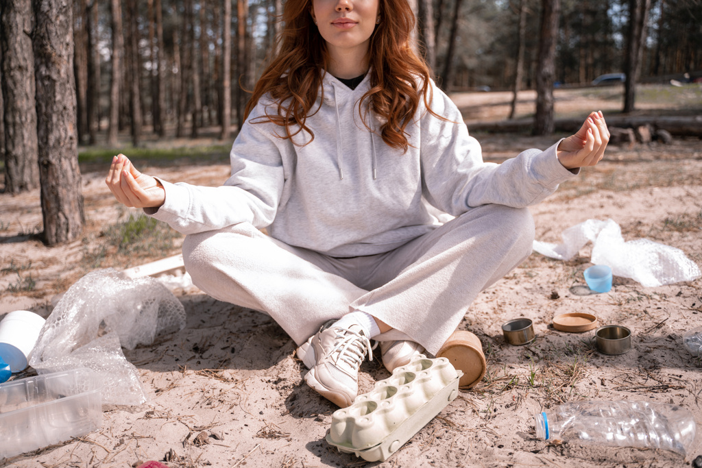 καλλιεργημένη άποψη της νεαρής γυναίκας με σταυρωμένα πόδια που κάθεται και διαλογίζεται κοντά σκουπίδια στο έδαφος - Φωτογραφία, εικόνα