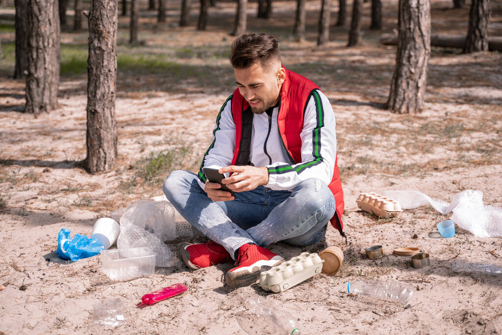 hombre alegre con las piernas cruzadas sentado y usando el teléfono inteligente cerca de la basura en el suelo - Foto, imagen