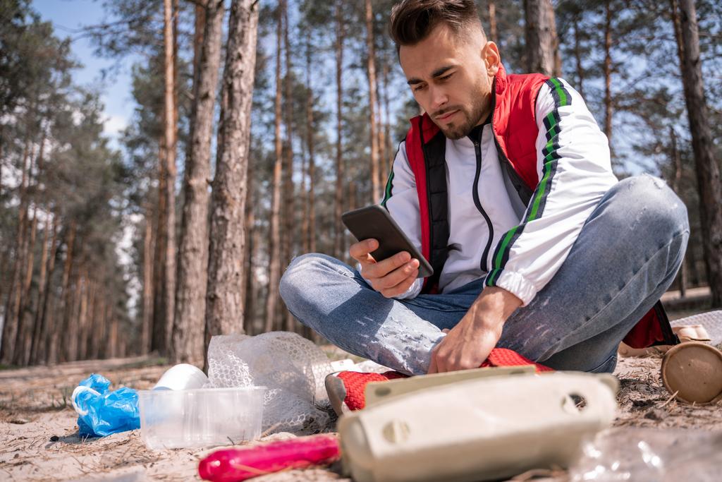 man with crossed legs sitting and using smartphone near trash on ground - Photo, Image