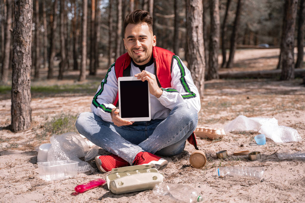 счастливый человек со скрещенными ногами сидит и держит цифровой планшет с пустым экраном рядом с мусором на земле - Фото, изображение