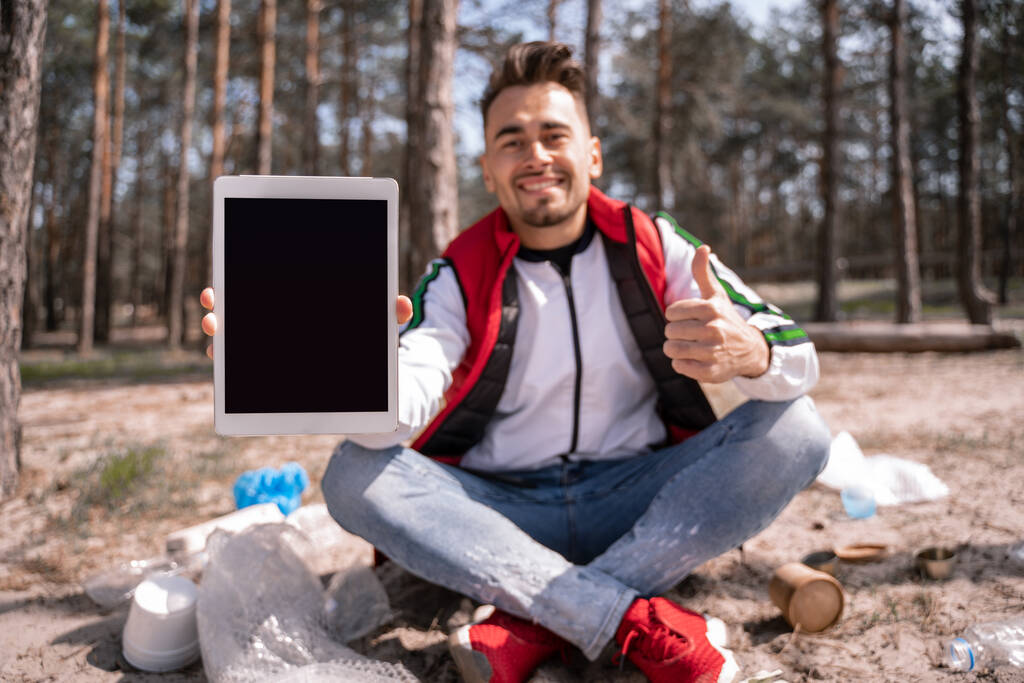 szczęśliwy człowiek ze skrzyżowanymi nogami trzymając cyfrowy tablet z pustym ekranem i pokazując kciuk w górę w pobliżu śmieci na ziemi - Zdjęcie, obraz