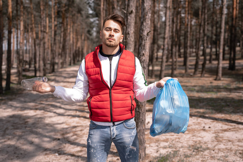 Молодой человек держит синий мешок для мусора и пластиковую бутылку в лесу  - Фото, изображение