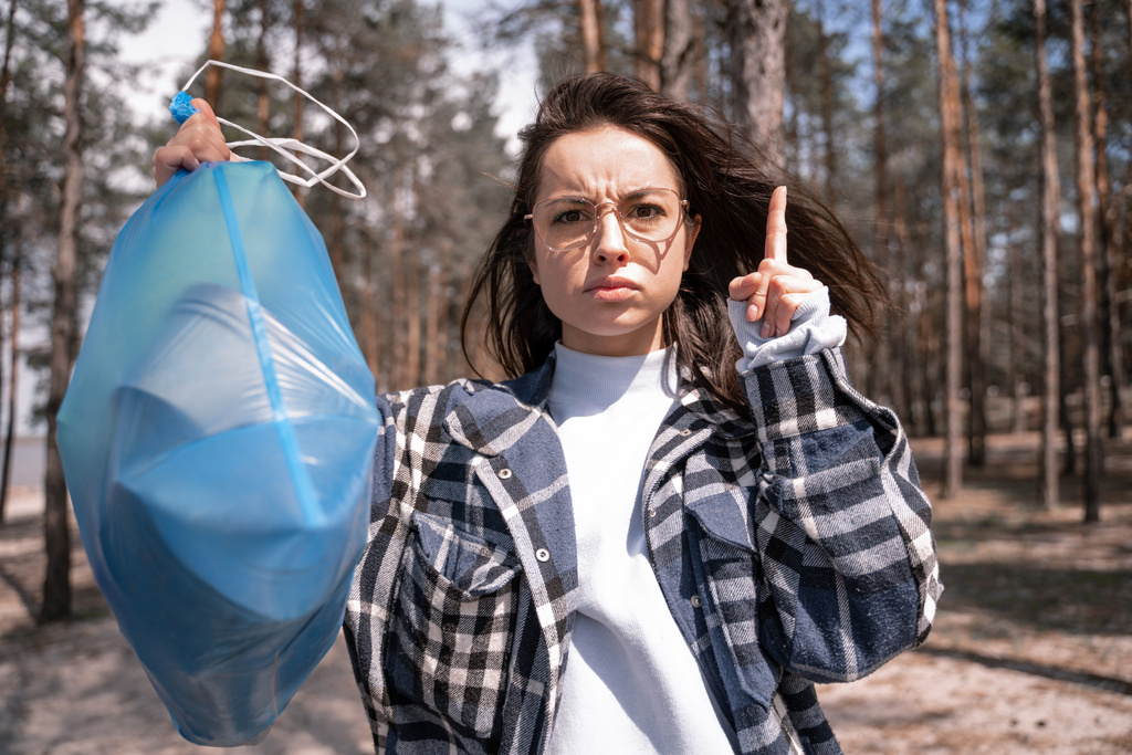 young woman warning while pointing with finger and holding blue trash bag in forest - Photo, Image
