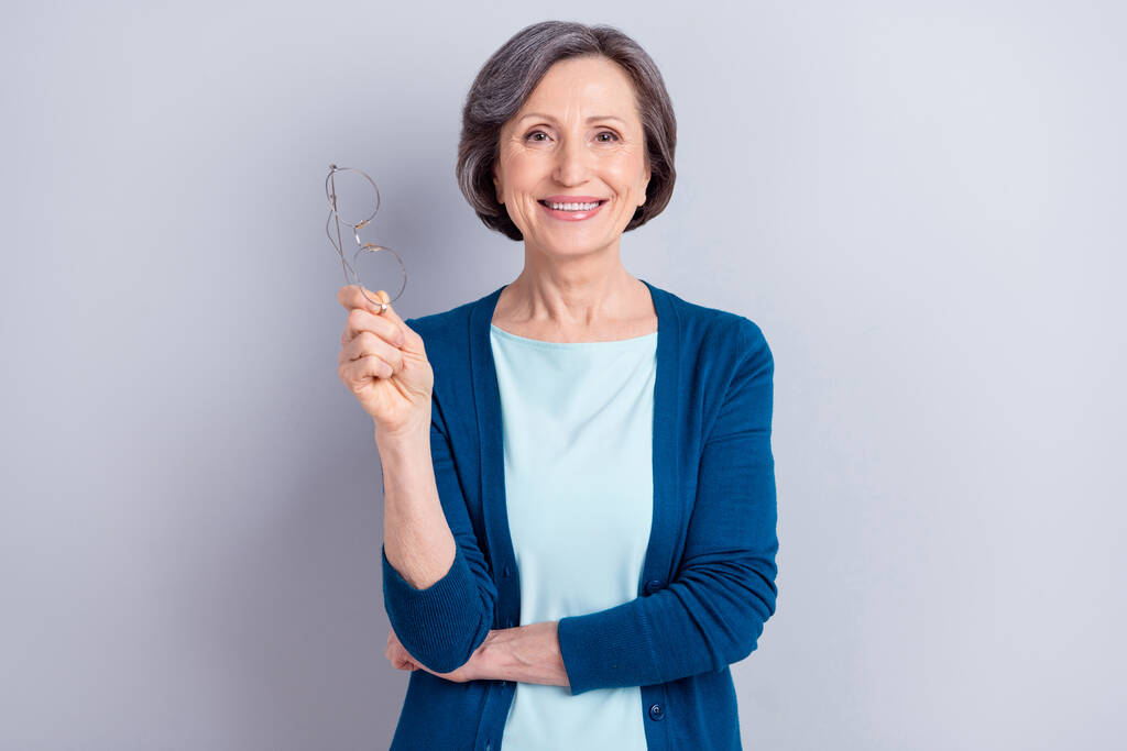 Фотопортрет щасливої літньої жінки, що тримає окуляри в блакитному кардигані, посміхаючись ізольовано на сірому кольоровому фоні
 - Фото, зображення