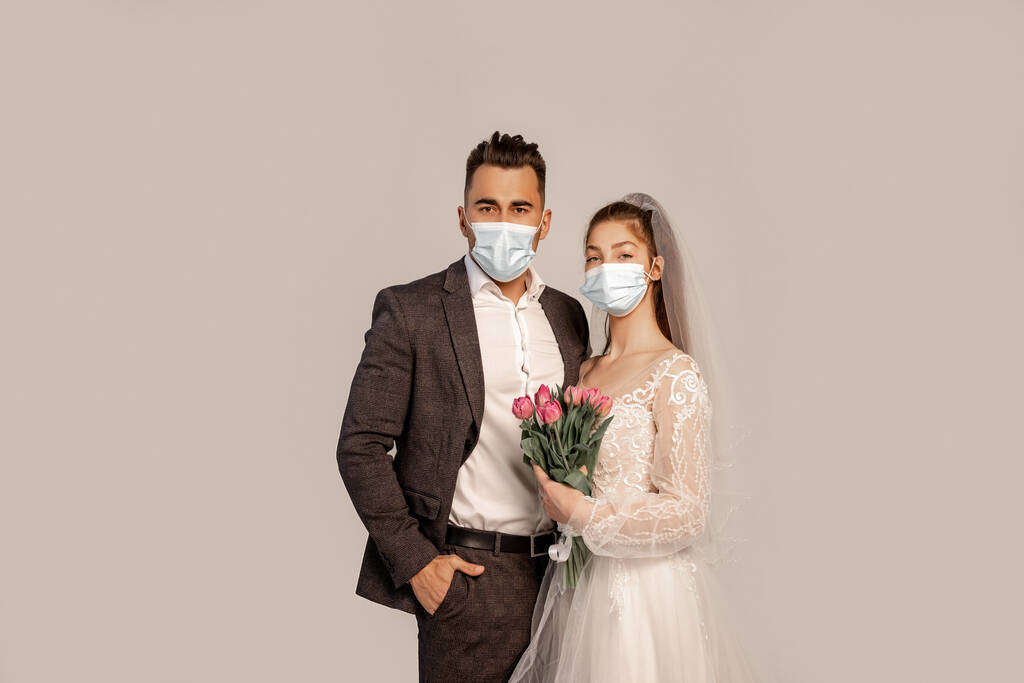 мужчина в медицинской маске, стоящий с рукой в кармане рядом с невестой с тюльпанами, изолированными на сером - Фото, изображение