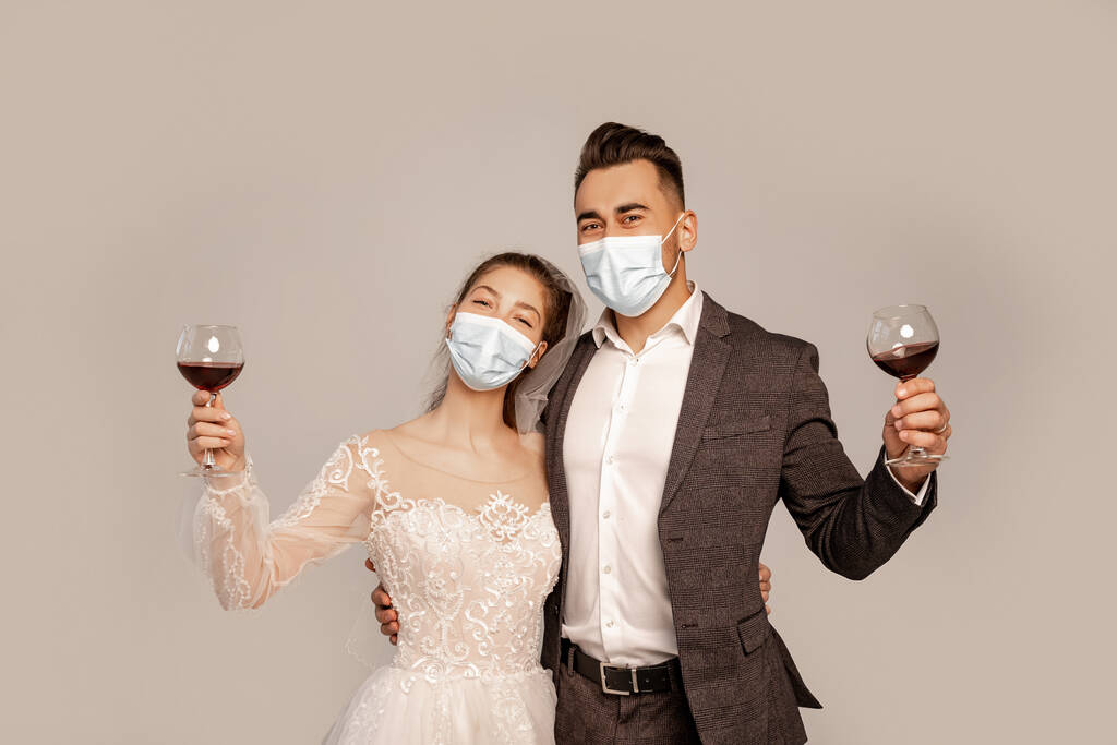 young newlyweds in medical masks holding glasses with red wine isolated on grey - Zdjęcie, obraz
