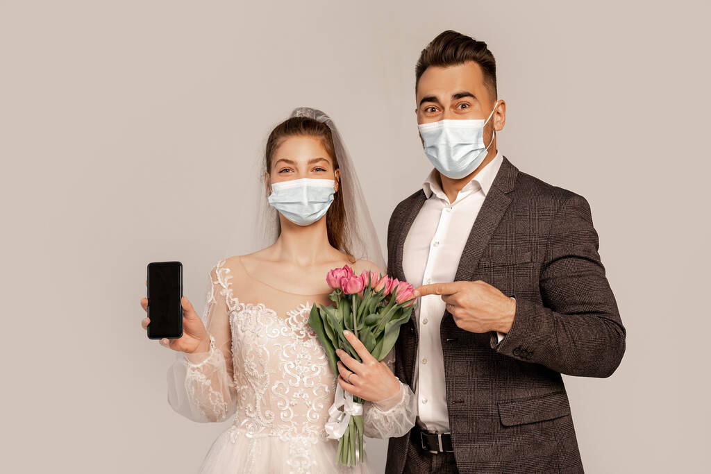 man in medical mask pointing at smartphone in hand of bride isolated on grey - 写真・画像