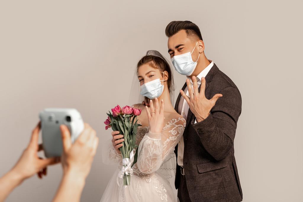 newlyweds in medical masks showing wedding rings near blurred photographer isolated on grey - Foto, afbeelding