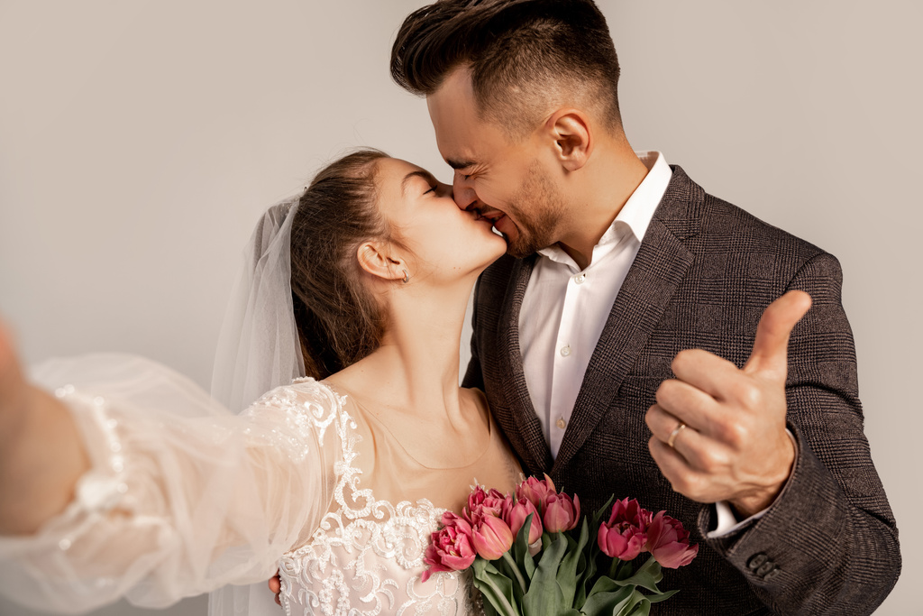 Mann zeigt Daumen nach oben, während er Braut auf grau mit fliederfarbenem Farbton küsst - Foto, Bild