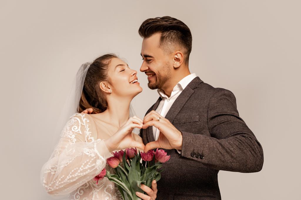 joyful newlyweds smiling at each other while showing heart symbol with hands isolated on grey - Φωτογραφία, εικόνα