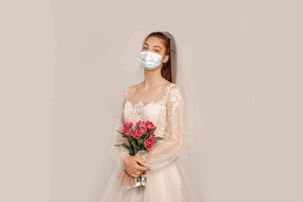 young bride in safety mask holding fresh tulips isolated on grey with lilac shade - Fotó, kép