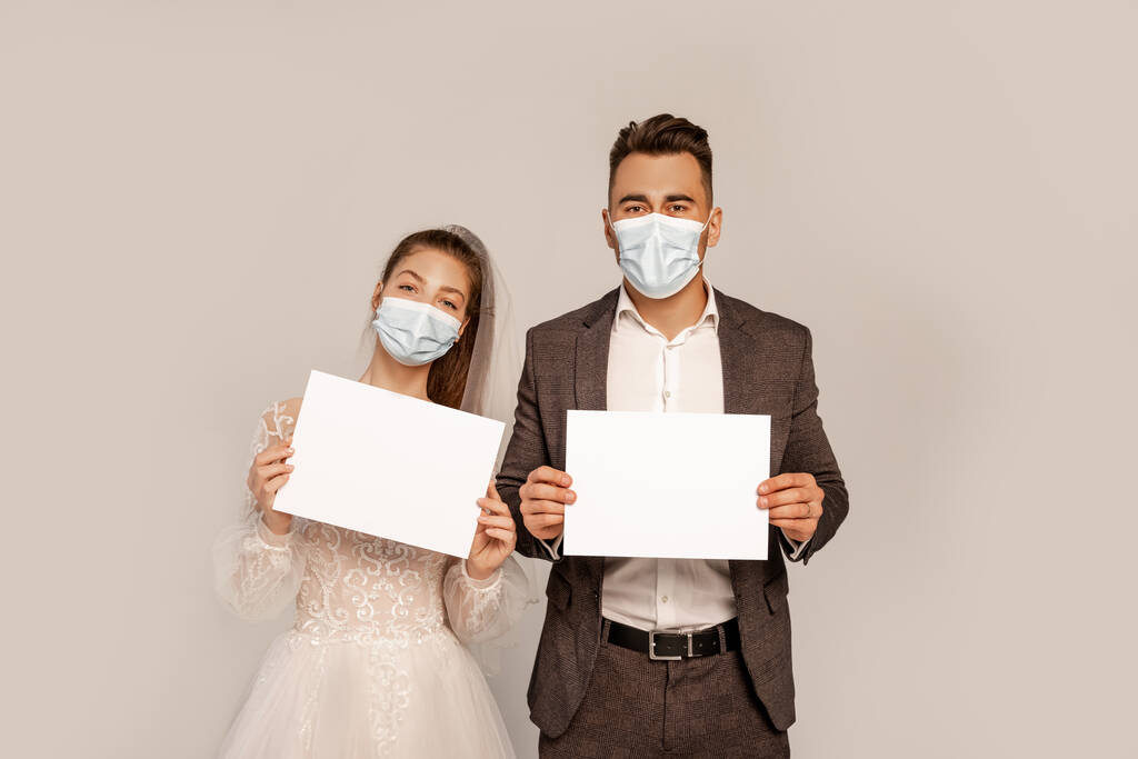 молодая пара в медицинских масках с пустыми плакатами, изолированными на серой - Фото, изображение