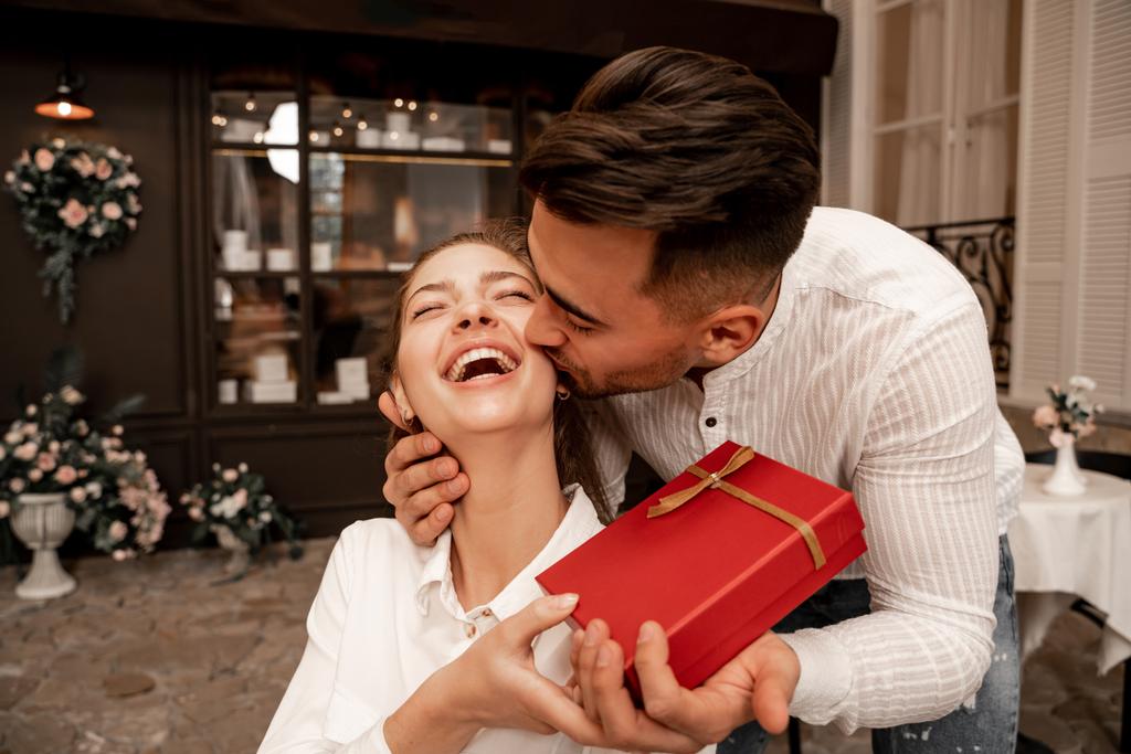 Молодой человек целует смеющуюся женщину, преподнося подарок в кафе - Фото, изображение