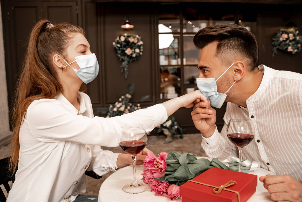young man in protective mask kissing hand of woman during date in cafe - Foto, imagen