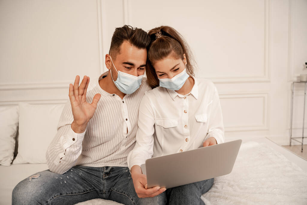 Mann mit medizinischer Maske winkt bei Videoanruf bei Freundin zu Hause - Foto, Bild
