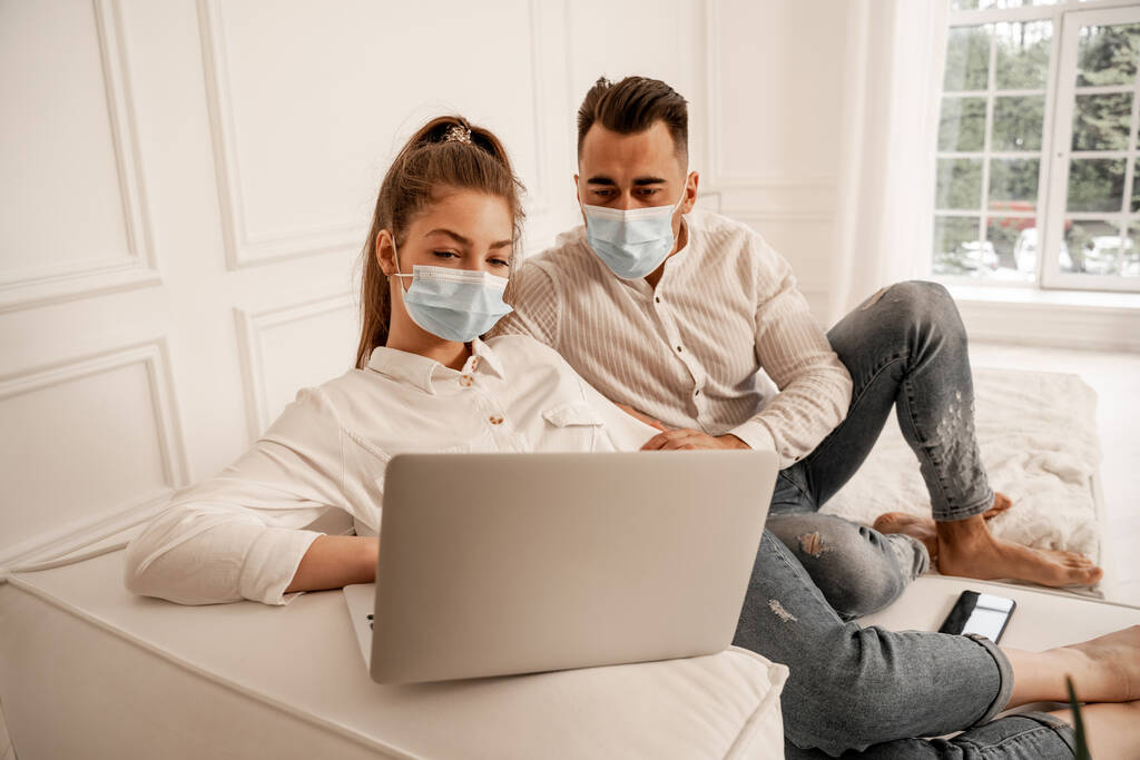 young couple in medical masks watching movie on laptop at home - Fotografie, Obrázek