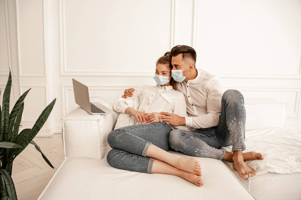 мужчина в медицинской маске обнимает подружку во время просмотра фильма на ноутбуке - Фото, изображение