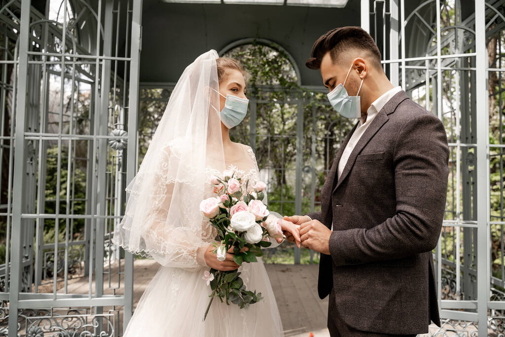 young man in medical mask putting wedding ring on finger of bride - Foto, afbeelding