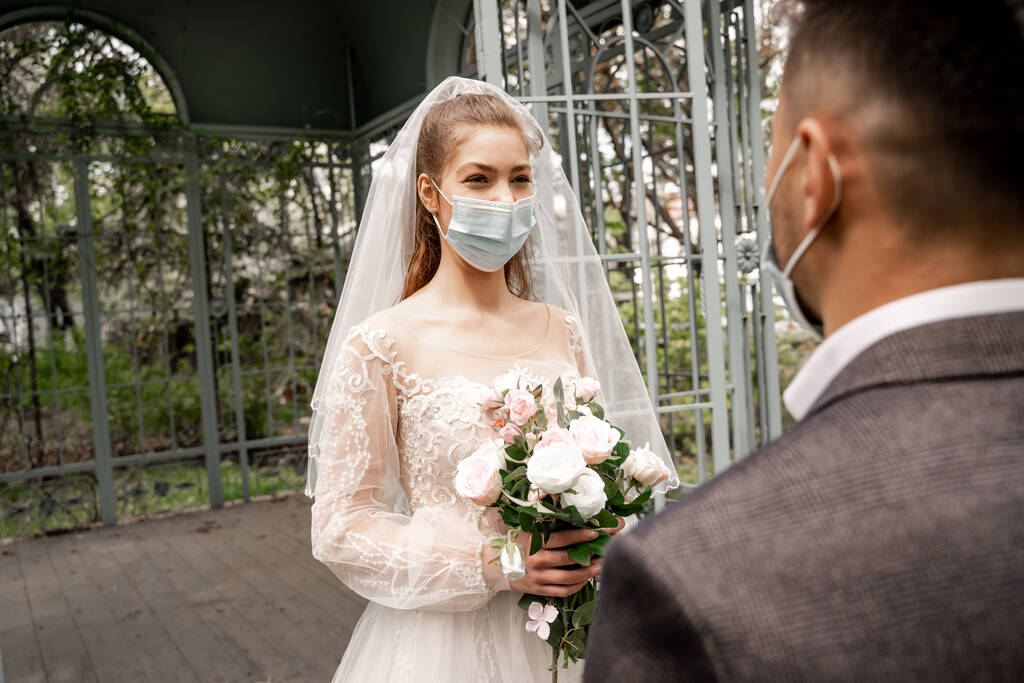 elegant bride in medical mask holding wedding bouquet near blurred groom in park - Fotoğraf, Görsel