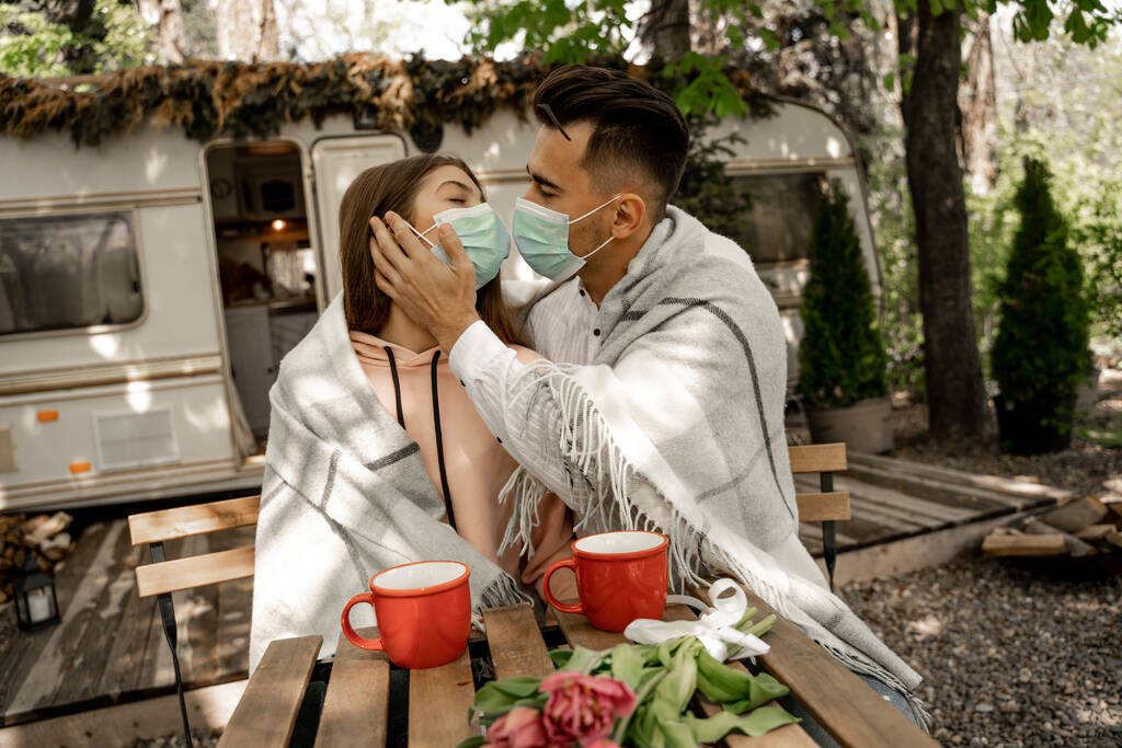 man in medical mask kissing girlfriend near cups of coffee outdoors - Zdjęcie, obraz