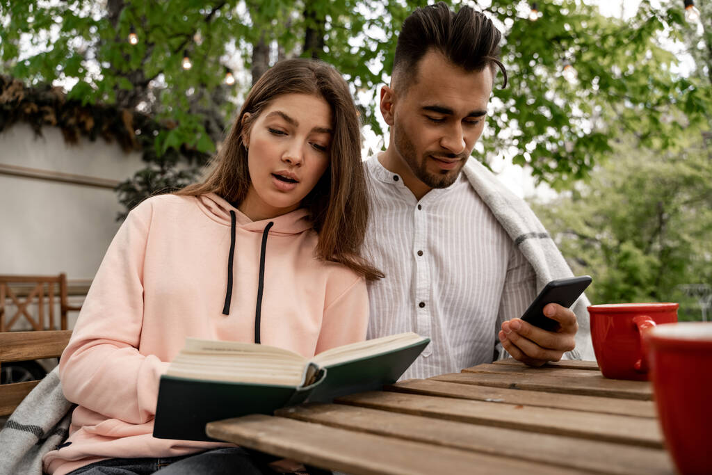 pretty woman reading book near man chatting on smartphone outdoors - Fotó, kép