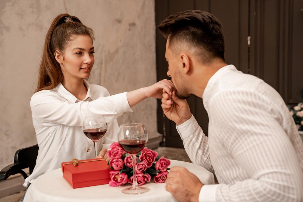 man kissing hand of pretty woman near wine glasses, present and bouquet in restaurant - 写真・画像