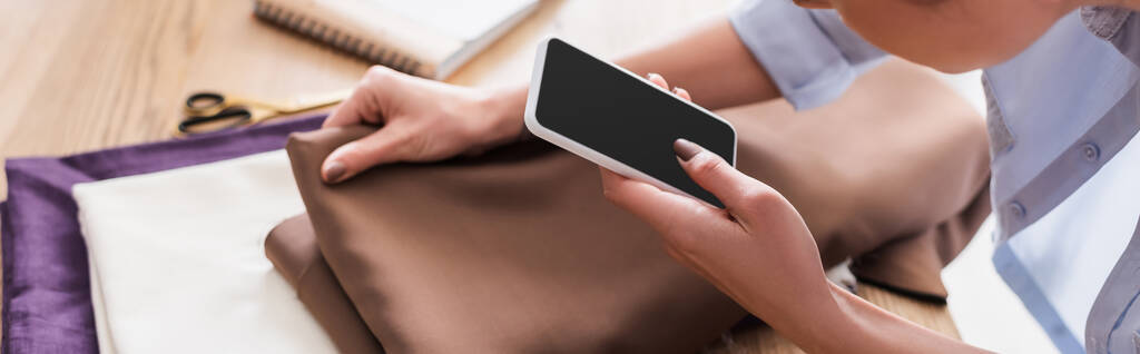 Widok szwaczki przy użyciu smartfona w pobliżu rozmytej tkaniny, baner  - Zdjęcie, obraz