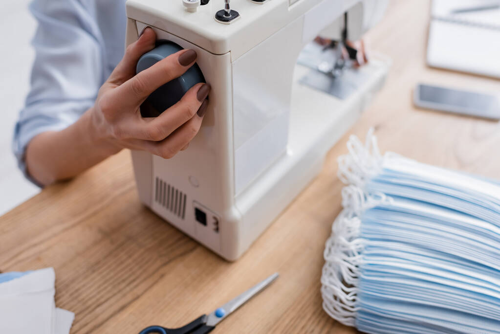 Vista recortada de la costurera que ajusta la máquina de coser cerca de máscaras médicas en la mesa  - Foto, imagen