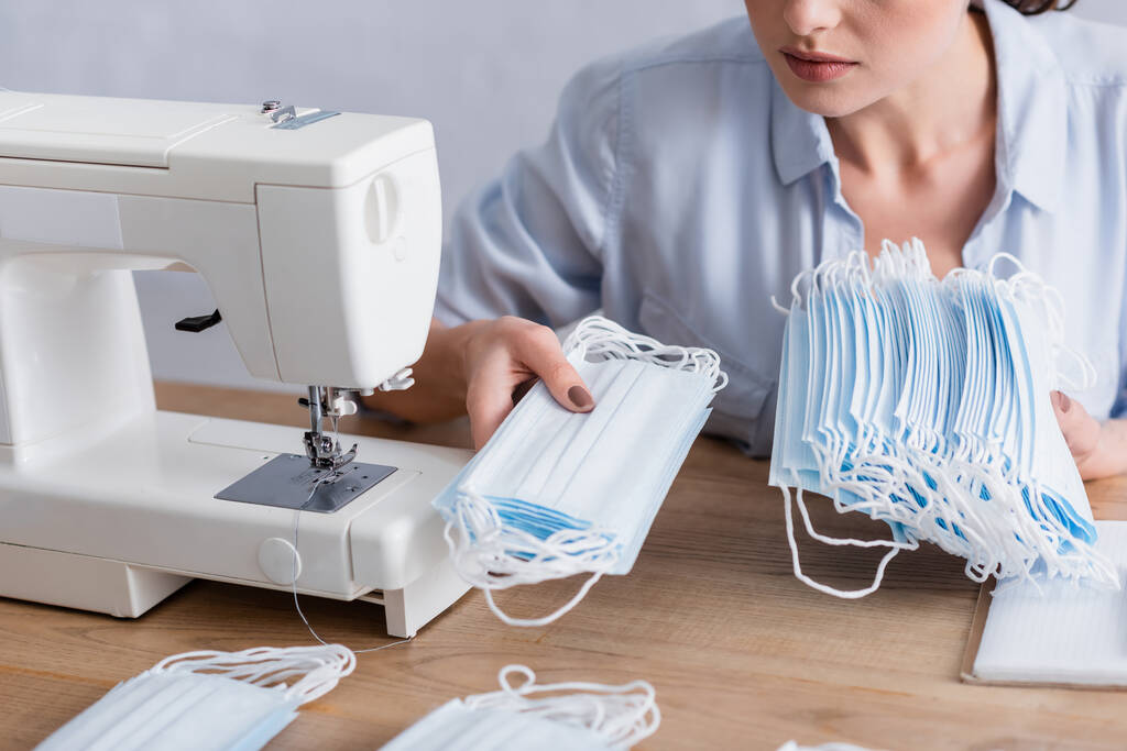 Обрезанный вид швеи, держащей медицинские маски возле ноутбука и швейной машинки  - Фото, изображение