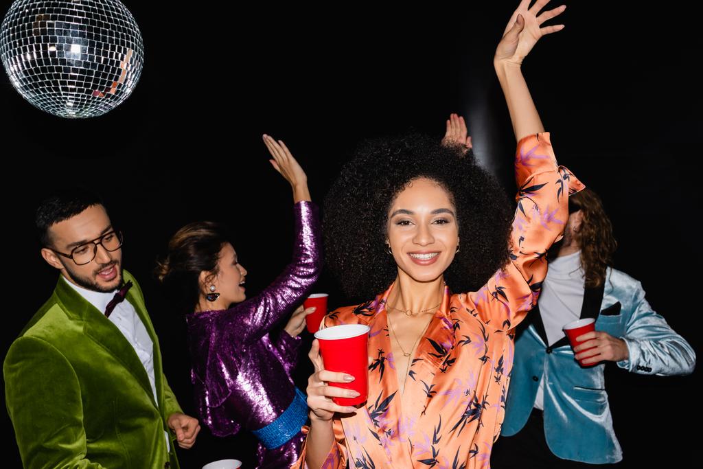 позитивная африканская американка танцует на вечеринке с многонациональными друзьями в разноцветной одежде на черном фоне - Фото, изображение