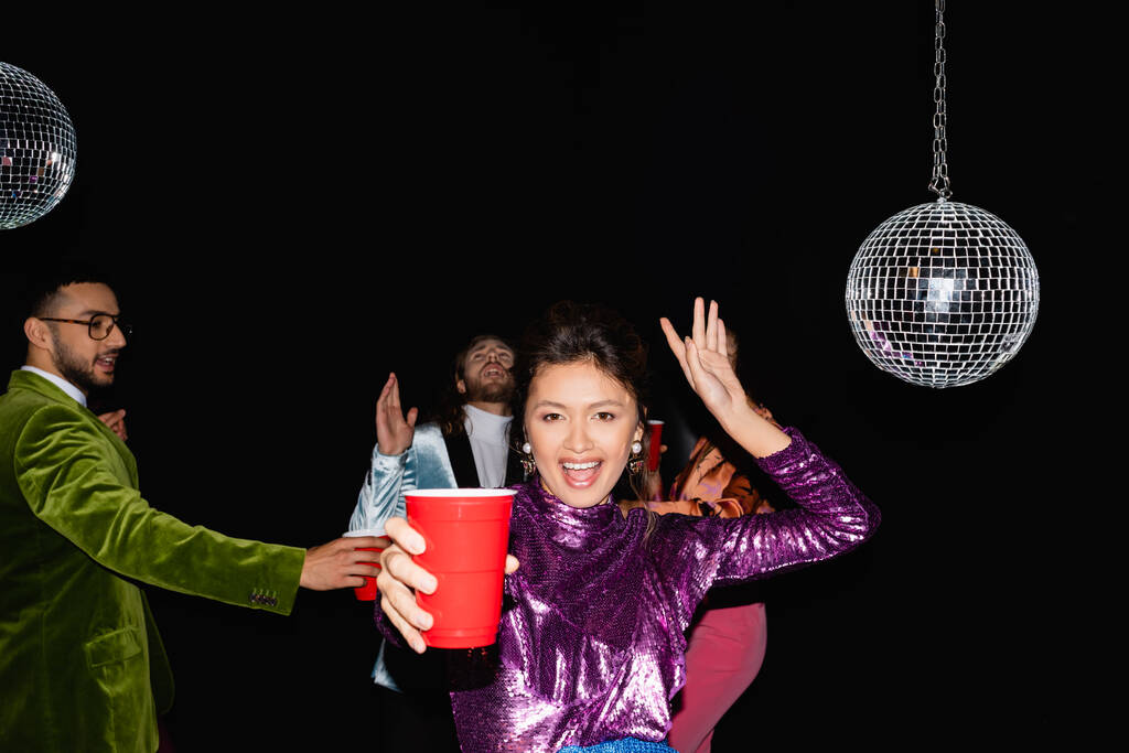 азиатка с пластиковой чашкой танцует рядом размытые многорасовые друзья в красочной одежде на черном фоне - Фото, изображение