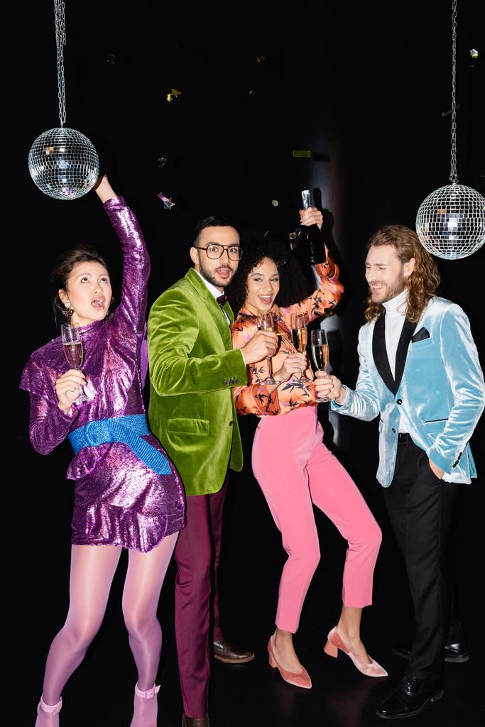 playful multiracial friends in colorful clothes with glasses of champagne in hands dancing on black background - Photo, Image