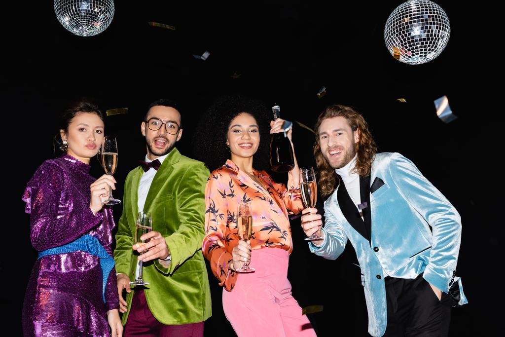 игривые многорасовые друзья в разноцветной одежде с бокалами шампанского, веселящиеся на черном фоне - Фото, изображение