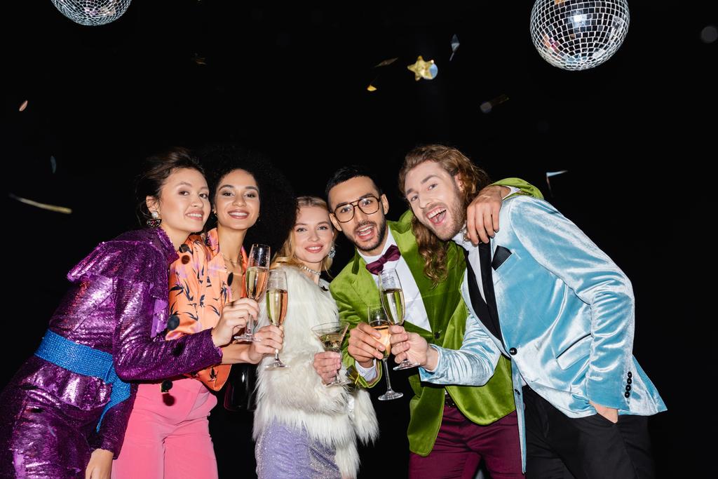 hravé multiraciální přátelé objímání s sklenicemi šampaňského na černém pozadí - Fotografie, Obrázek