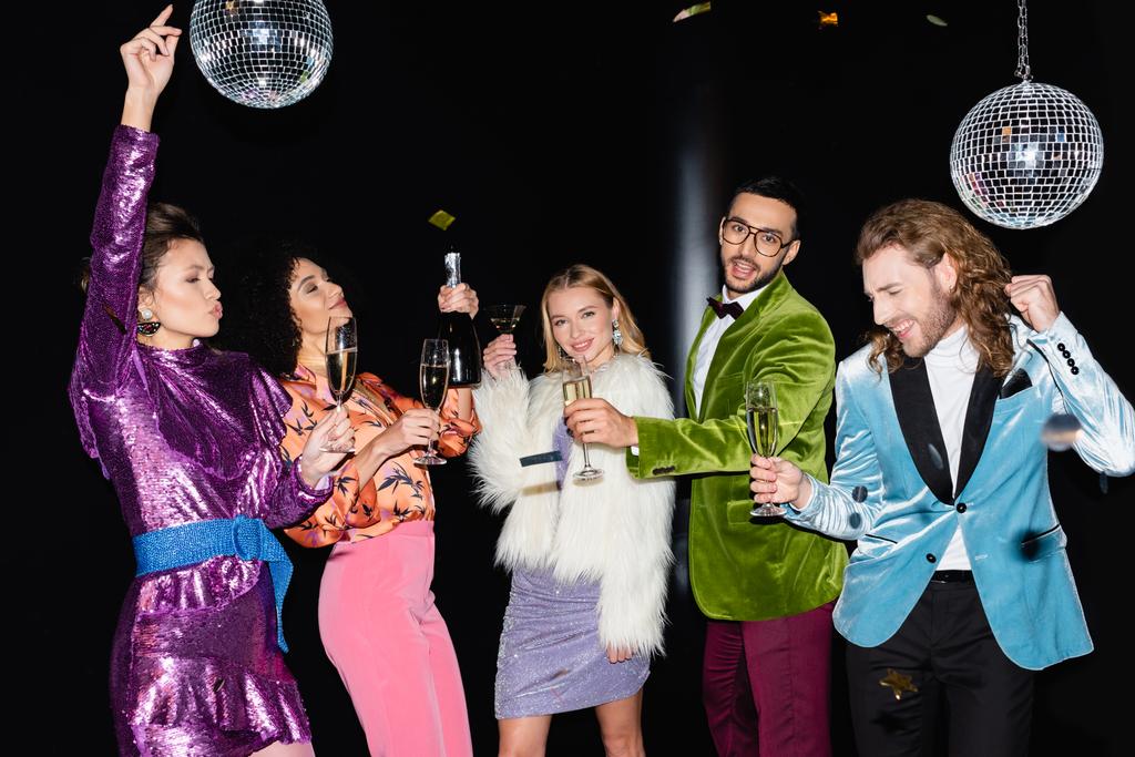 amici multirazziali positivi in vestiti colorati che tengono bicchieri di champagne e ballano su sfondo nero - Foto, immagini