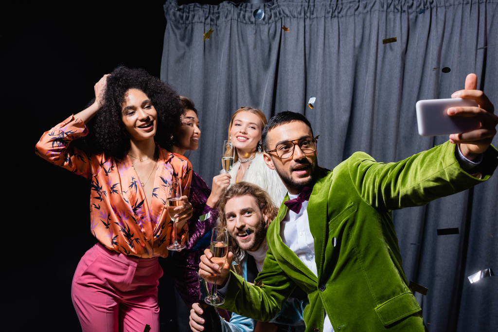 amigos inter-raciais elegantes em roupas brilhantes tomando selfie no smartphone perto de cortina cinza no fundo preto - Foto, Imagem