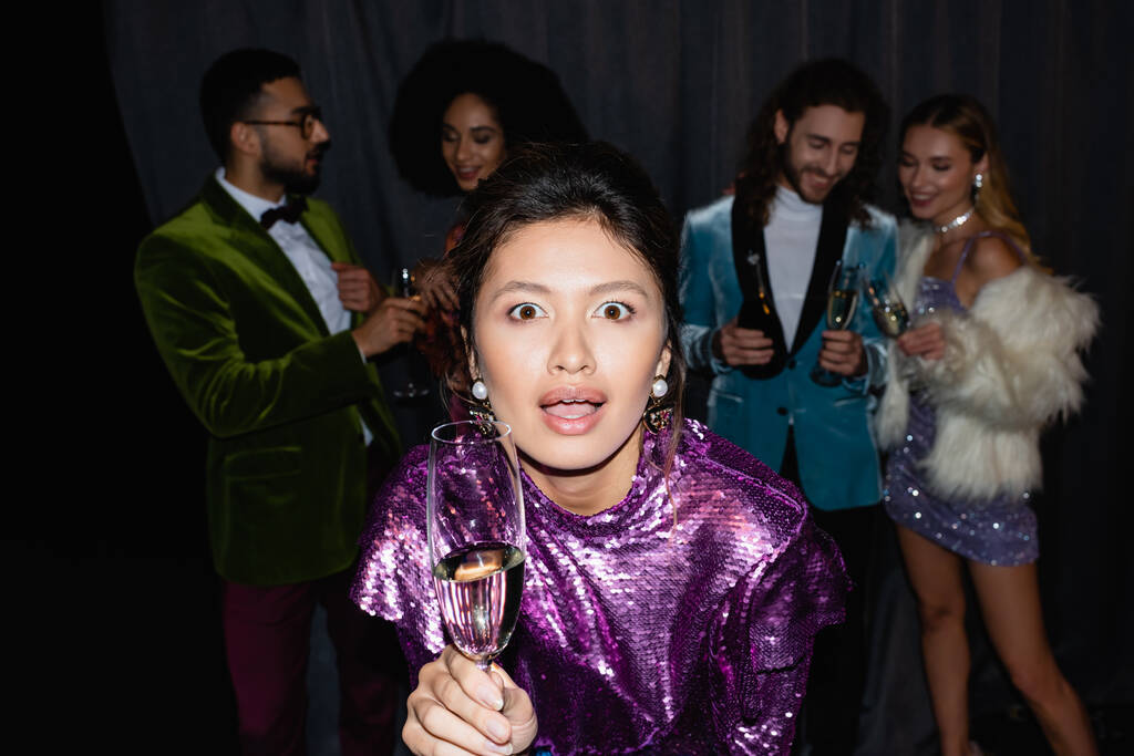 asiático joven adulto mujer mirando a cámara y celebración de la copa de champán cerca borrosa interracial amigos en gris fondo - Foto, imagen