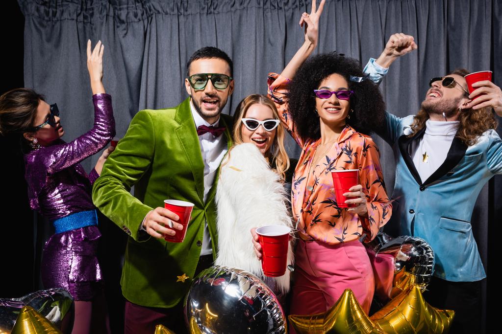 θετικοί διαφυλετικοί φίλοι με πολύχρωμα ρούχα και γυαλιά ηλίου χορεύουν κοντά σε γκρι κουρτίνα σε μαύρο φόντο - Φωτογραφία, εικόνα