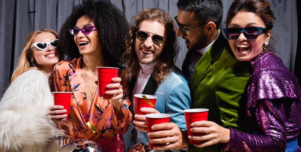 amigos interraciales juguetones en gafas de sol celebrando con copas de plástico sobre fondo gris, pancarta - Foto, imagen