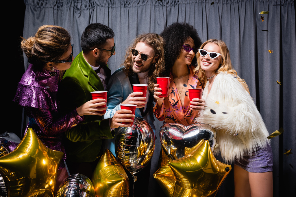 felices amigos interracial en gafas de sol celebrando con copas de plástico cerca de cortina gris sobre fondo negro - Foto, Imagen