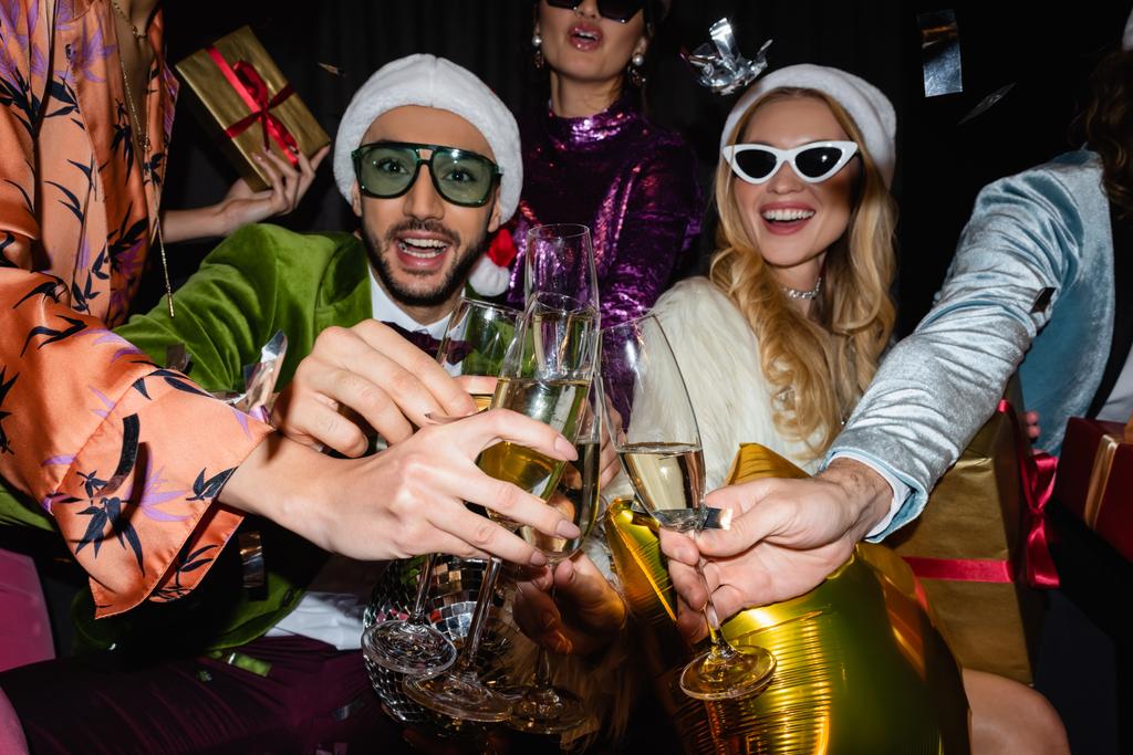 smiling interracial friends in santa hats and sunglasses clinking with glasses of champagne on black background - Photo, Image