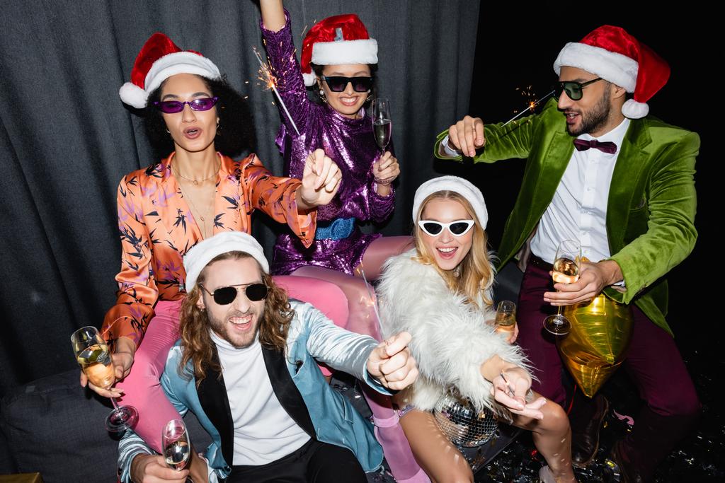 smiling interracial friends in santa hats and sunglasses celebrating new year near grey curtain on black background - Photo, Image