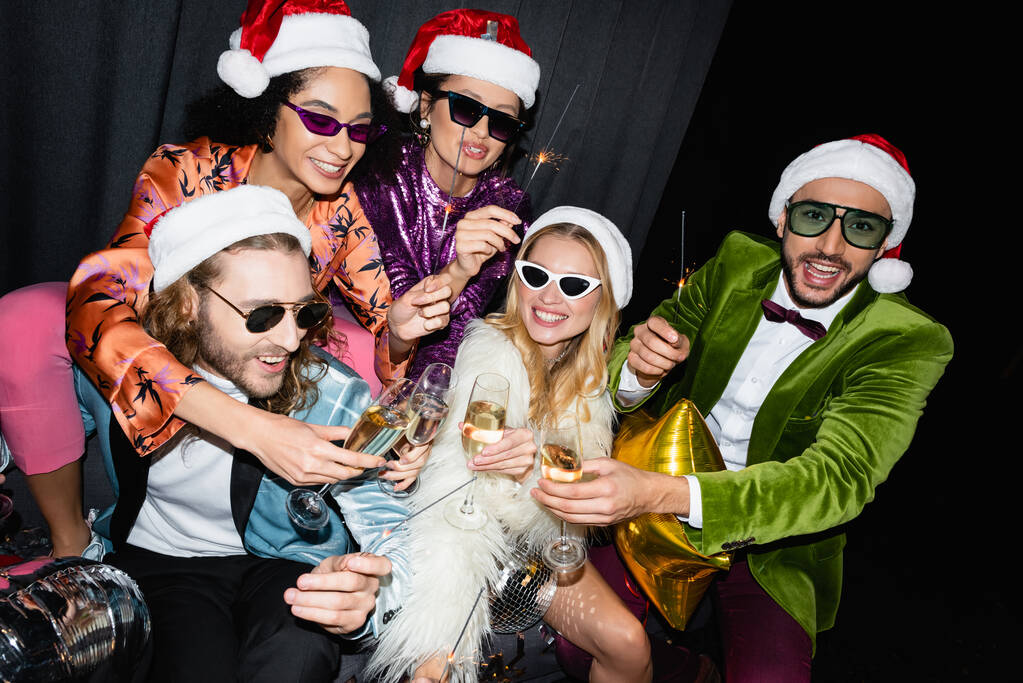 игривые межрасовые друзья в шляпах Санта-Клауса празднуют Новый год возле серого занавеса на черном фоне - Фото, изображение