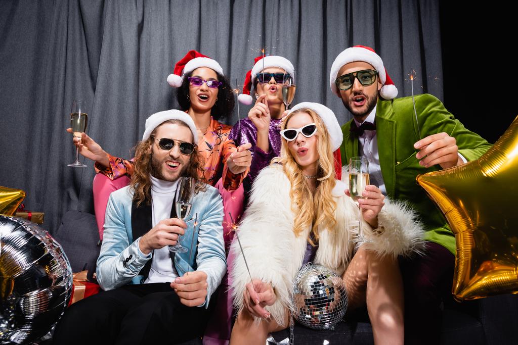 межрасовые друзья в шляпах Санта-Клауса празднуют Новый год возле серого занавеса на черном фоне - Фото, изображение